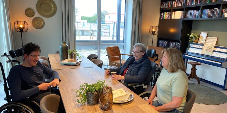 Jongen in rolstoel een oudere en een mevrouw van middelbare leeftijd praten met elkaar aan tafel in een ontmoetingsruimte met boekenkast en piano