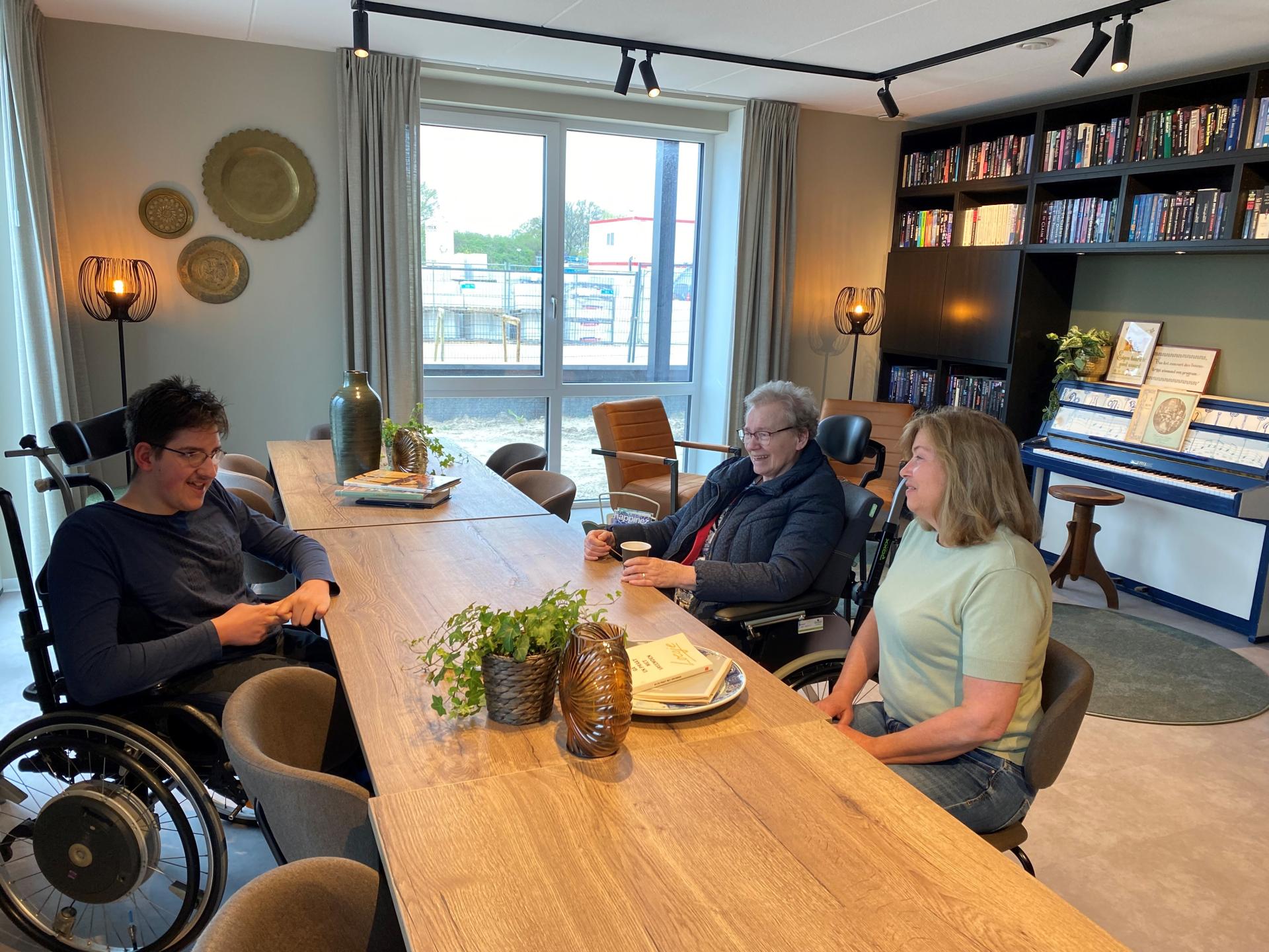 Jongen in rolstoel een oudere en een mevrouw van middelbare leeftijd praten met elkaar aan tafel in een ontmoetingsruimte met boekenkast en piano